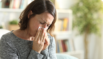 Woman sitting indoors holding a tissue to her nose, appearing to sneeze