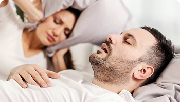Man sleeping on his back with mouth open, appearing to snore, while a woman beside him covers her ears with a pillow