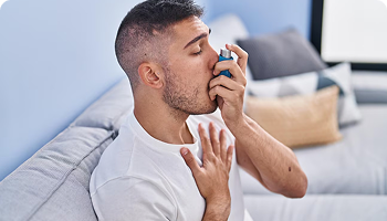 A young man sitting on a couch using an inhaler, with one hand on his chest, indicating difficulty breathing or an asthma attack.