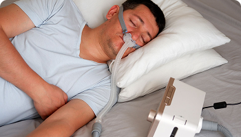 Man sleeping in bed while using a CPAP machine with a nasal mask, typically used to treat sleep apnea or other respiratory conditions.
