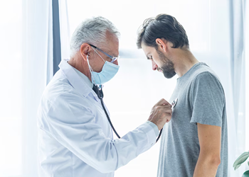 A doctor wearing a face mask uses a stethoscope to examine the chest of a male patient during a medical check-up in a brightly lit room.